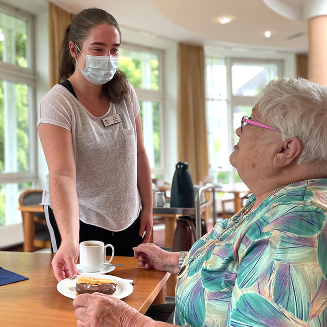 Eine Auszubildende der Hauswirtschaft serviert einer Bewohnerin Kaffee und Kuchen. Maria serviert der Bewohnerin Kaffee und Kuchen.